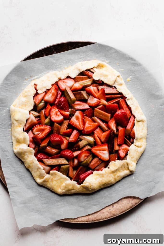 The assembled strawberry rhubarb galette, meticulously brushed with an egg wash and sprinkled with coarse sugar, poised on a baking pan and ready for the oven.
