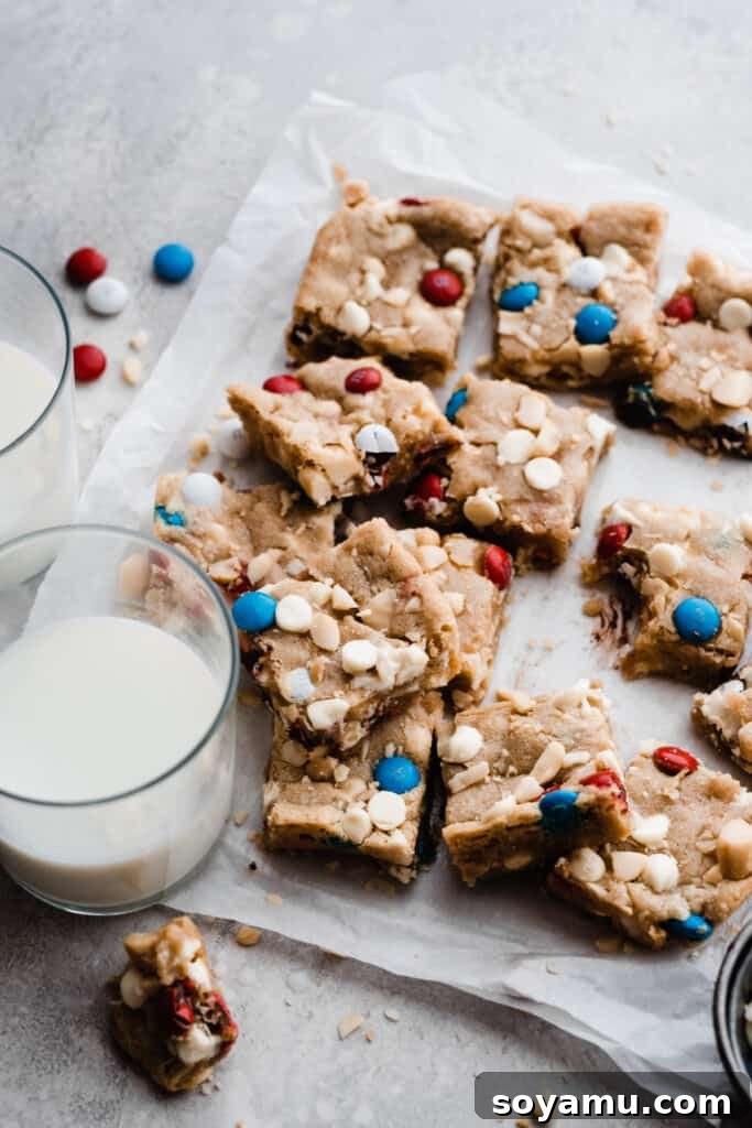 Vibrant M&M Fireworks Cookie Bars 6 Stacked cookie bars on parchment paper with two glasses of milk nearby, showcasing their delicious appeal.