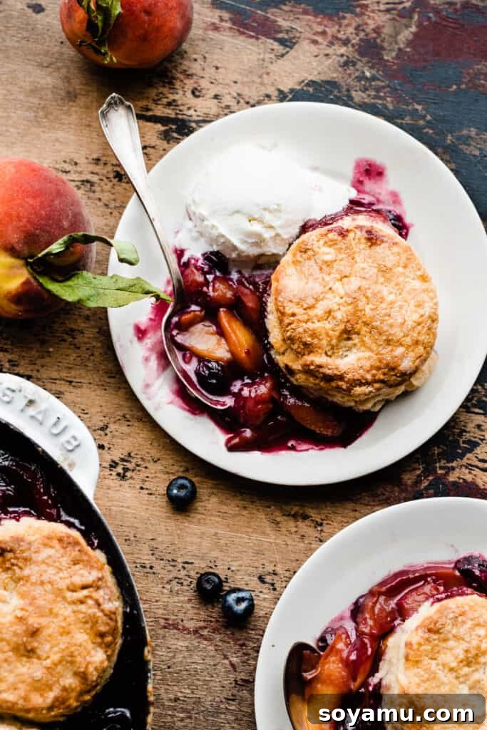 Two plates of vibrant blueberry peach cobbler, garnished with fresh blueberries and peach slices, elegantly presented on a rustic wooden surface, highlighting the golden biscuit topping and rich fruit filling.