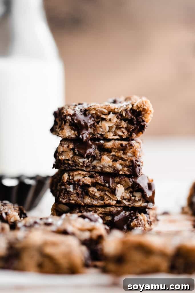 Chewy Brown Butter Chocolate Chunk Oatmeal Bars 2 A stack of golden-brown oatmeal chocolate chunk cookie bars, showcasing melty pools of chocolate and a sprinkle of flaky sea salt. A bottle of milk is visible in the soft-focus background.