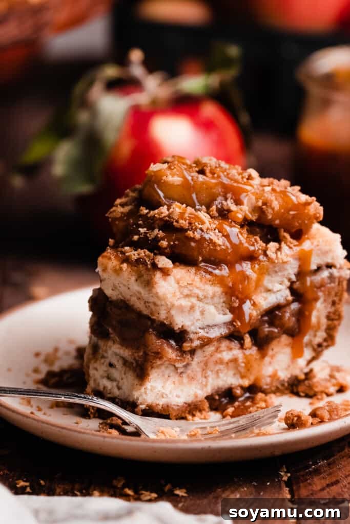 Decadent Salted Caramel Apple Pie Cheesecake 3 A close-up of a slice of apple cheesecake on a white plate, showing thick caramelized apples, streusel topping, and a rich caramel drizzle. A bite is missing from the slice.