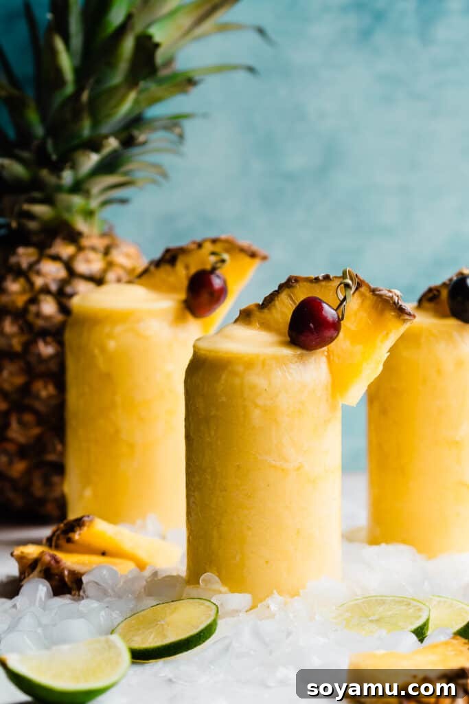 Paradise Frost 7 Three glasses of the frozen pineapple piña coladas with a whole pineapple nearby against a bright blue backdrop, emphasizing tropical freshness.