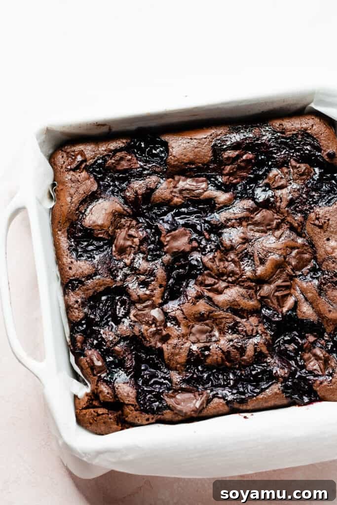 A baked pan of delectable Black Forest Brownies, showcasing a stunning marbled swirl of deep, dark chocolate brownie and vibrant red cherry sauce on its surface. The brownies appear perfectly set and ready to be cooled and sliced, promising a delicious and visually appealing dessert.
