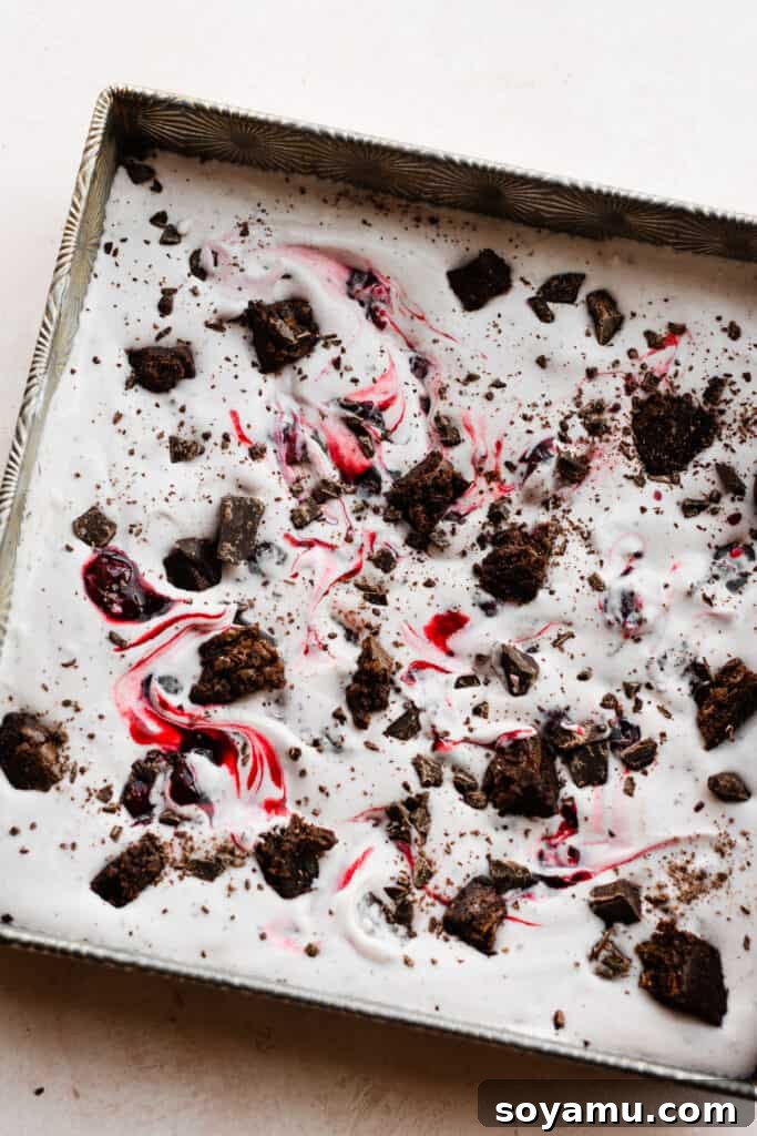 Effortless Black Forest Bliss 6 A square pan filled with the first layer of Black Forest Ice Cream base, generously topped with a vibrant swirl of cherry sauce, fudgy brownie pieces, and glistening chocolate chunks.