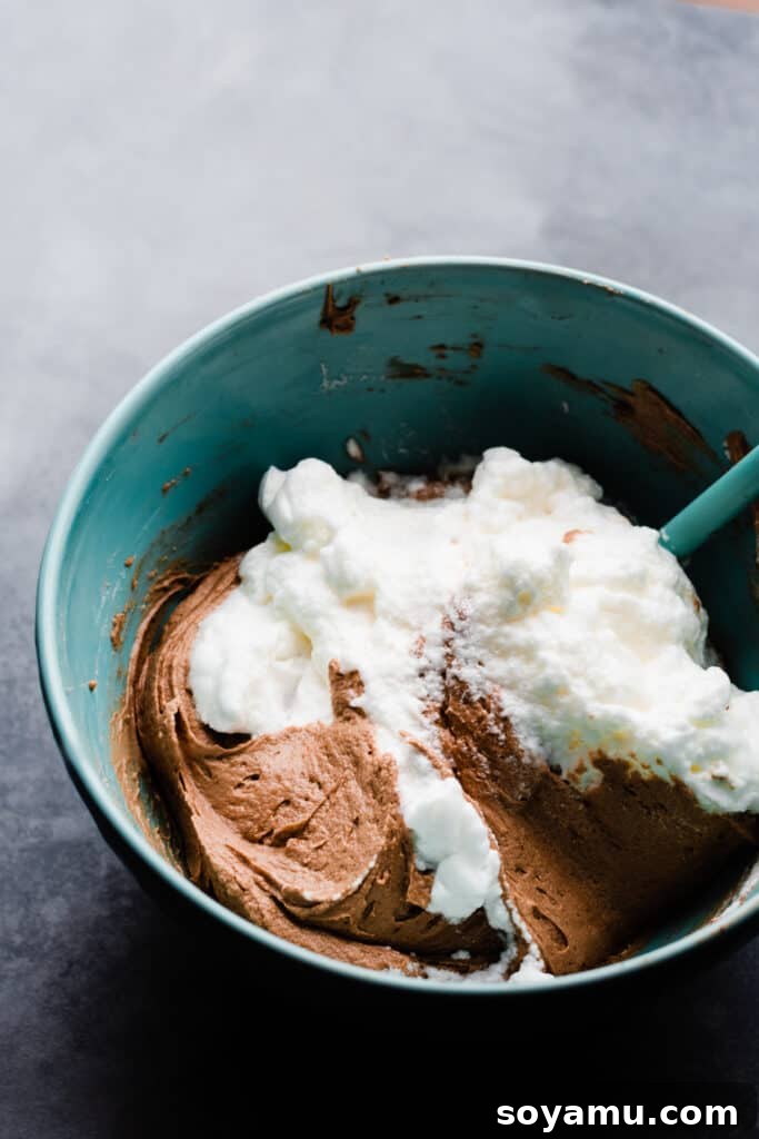 A spatula gently folding fluffy beaten egg whites into the dark, rich chocolate cake batter, illustrating the delicate technique.