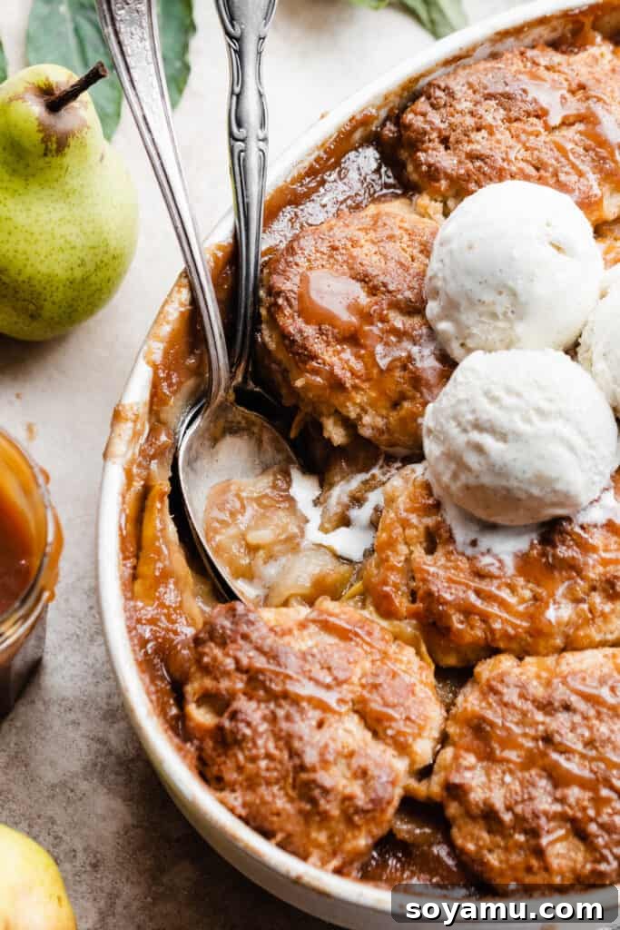 Caramel Sea Salt Pear Cobbler 10 A serving spoon expertly digs into the warm, luscious pear cobbler, revealing the tender fruit filling and flaky biscuit topping, with creamy vanilla ice cream melting into the mixture.
