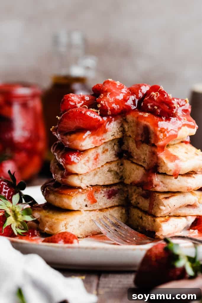 Fluffy Strawberry Pancakes with Speedy Strawberry Compote 2 A tall stack of fluffy strawberry pancakes, with a wedge missing, revealing the tender, berry-filled interior. They are generously drizzled with bright red strawberry compote.