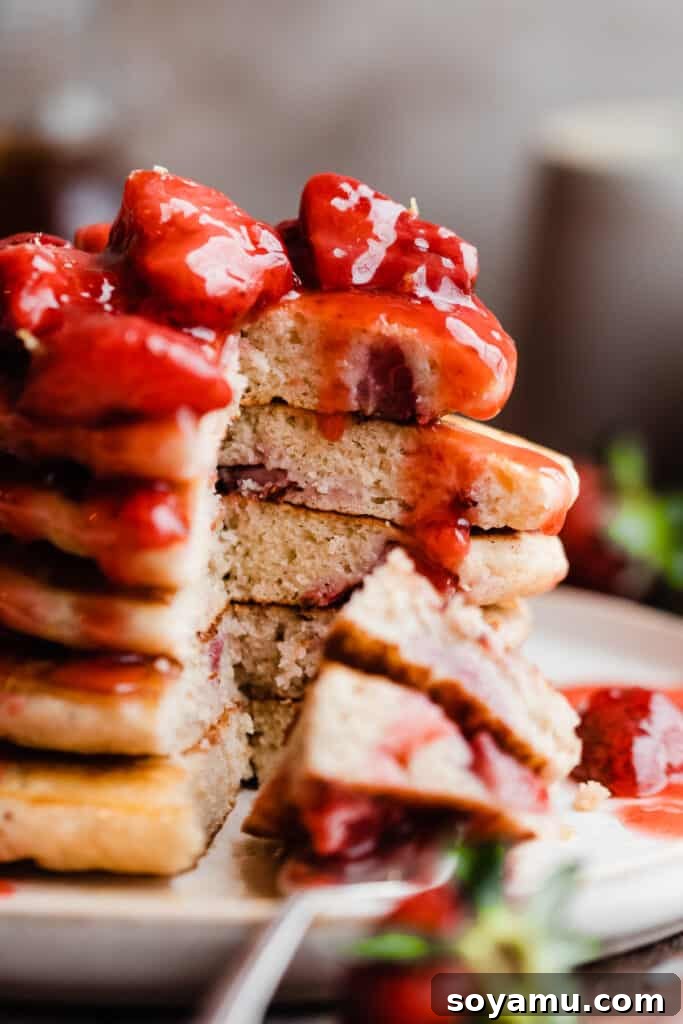 Fluffy Strawberry Pancakes with Speedy Strawberry Compote 11 A close-up shot of a stack of fluffy strawberry pancakes with a bite taken out, showing the light, airy texture and embedded strawberry pieces, topped with compote.