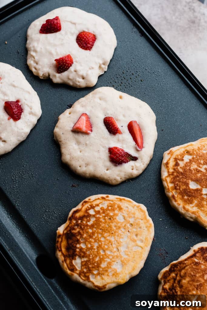 Fluffy Strawberry Pancakes with Speedy Strawberry Compote 7 Several strawberry pancakes cooking on a hot griddle, showing the bubbling batter and golden edges.
