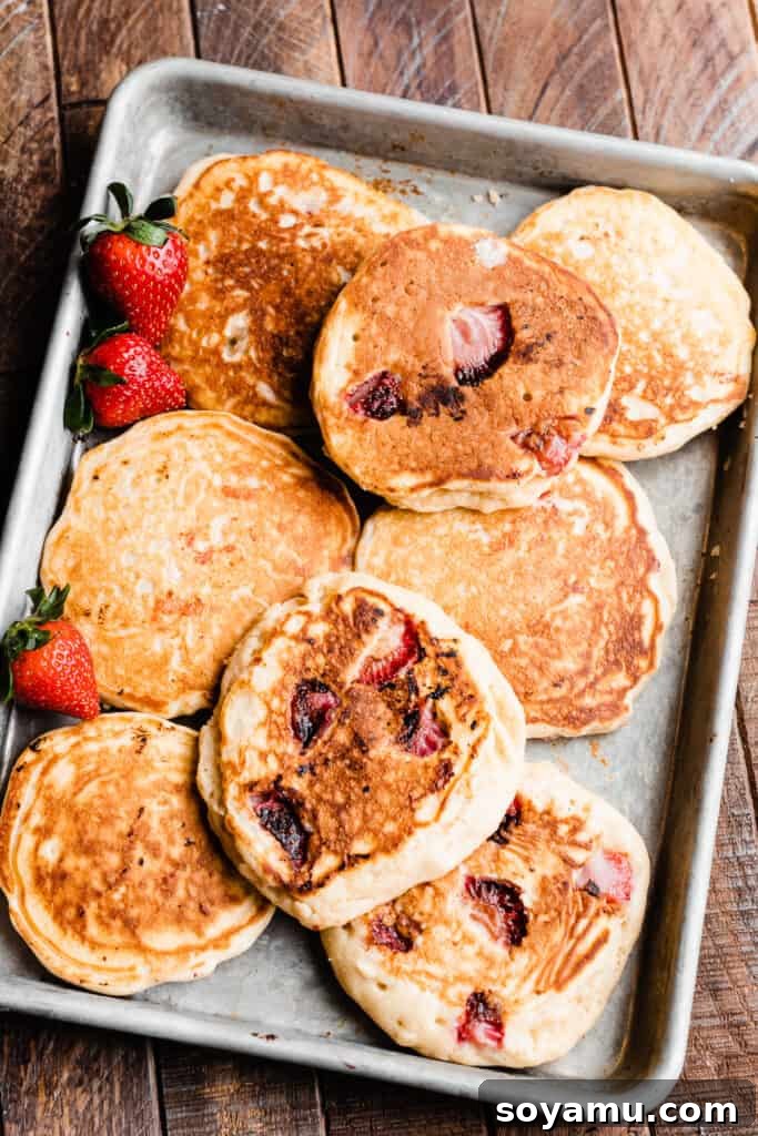 Fluffy Strawberry Pancakes with Speedy Strawberry Compote 8 Freshly cooked strawberry pancakes neatly arranged on a baking sheet, keeping warm before serving.