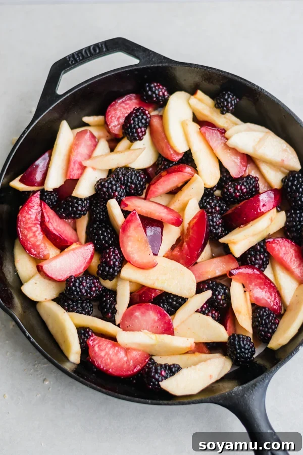 Mixed apples, plums, and blackberries generously spread out in a seasoned cast iron pan, ready for the crumble topping.