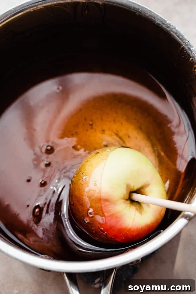 A fresh apple on a wooden skewer being carefully dipped into a pot of warm, red candy mixture, ensuring an even coating.