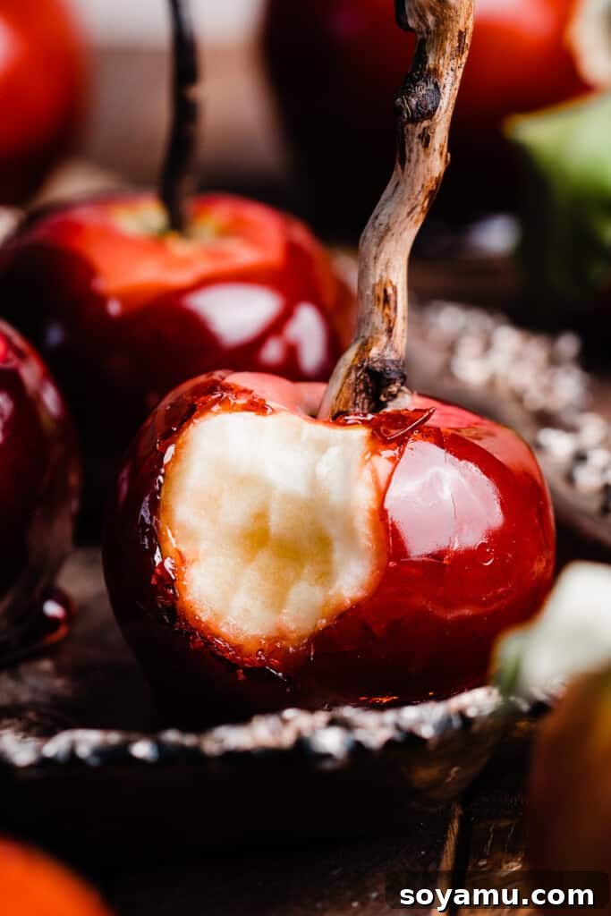 A close-up of a vibrant red toffee apple with a bite taken out, showcasing the crisp apple underneath its hard candy shell, with a wooden skewer.