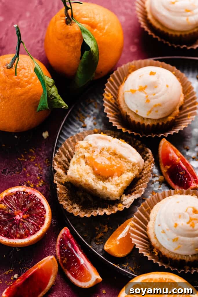 A beautifully arranged plate of frosted orange cupcakes, with one cupcake expertly halved to showcase its generous orange curd filling and soft, tender crumb.