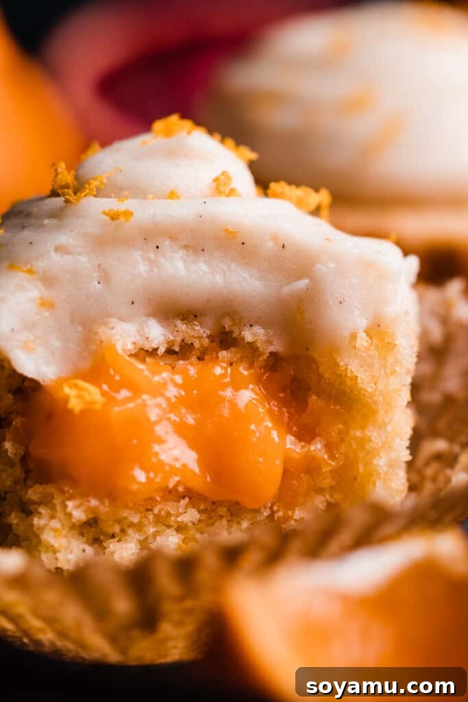 A close-up shot of an orange cupcake with a bite taken out, clearly showcasing the lush orange curd filling and the soft texture of the cake underneath the smooth frosting.