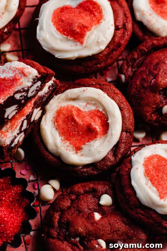 Decadent Red Velvet Cookies with Cream Cheese Frosting 3 A trio of freshly baked red velvet cookies, each adorned with creamy white cream cheese frosting and a vibrant red heart-shaped sprinkle design in the center, showcased beautifully.