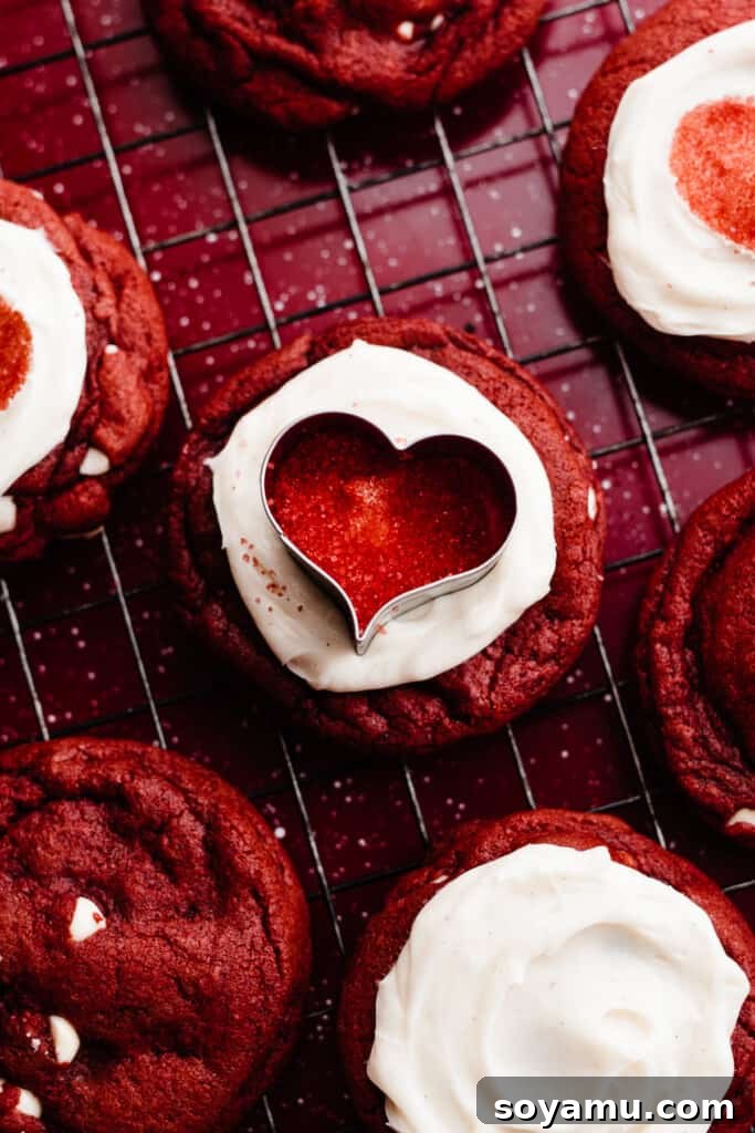 Decadent Red Velvet Cookies with Cream Cheese Frosting 8 A close-up of a hand gently decorating a frosted red velvet cookie with red sprinkles, using a small heart-shaped cookie cutter as a stencil for a precise design.