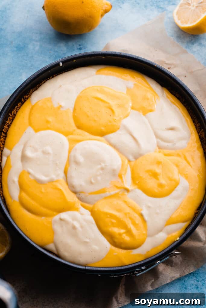 Alternating dollops of light lemon cheesecake batter and darker lemon curd cheesecake batter beautifully arranged in the springform pan over the crust, ready for swirling.
