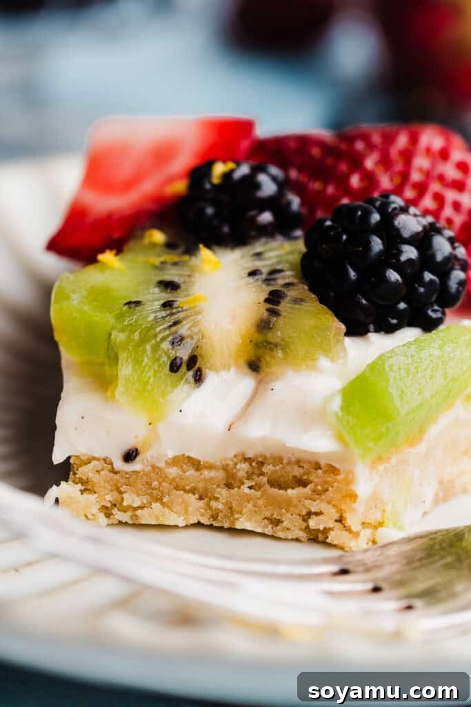 A tantalizing close-up of a fruit pizza slice on a plate, with a small bite taken, revealing the delightful chewy texture of the sugar cookie base beneath the creamy frosting and colorful fruit.