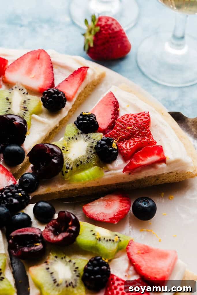 A delectable close-up of a single slice of Fruit Pizza, showing off its generous layer of cream cheese frosting and a colorful mosaic of fresh fruit, artfully arranged on a marble platter.