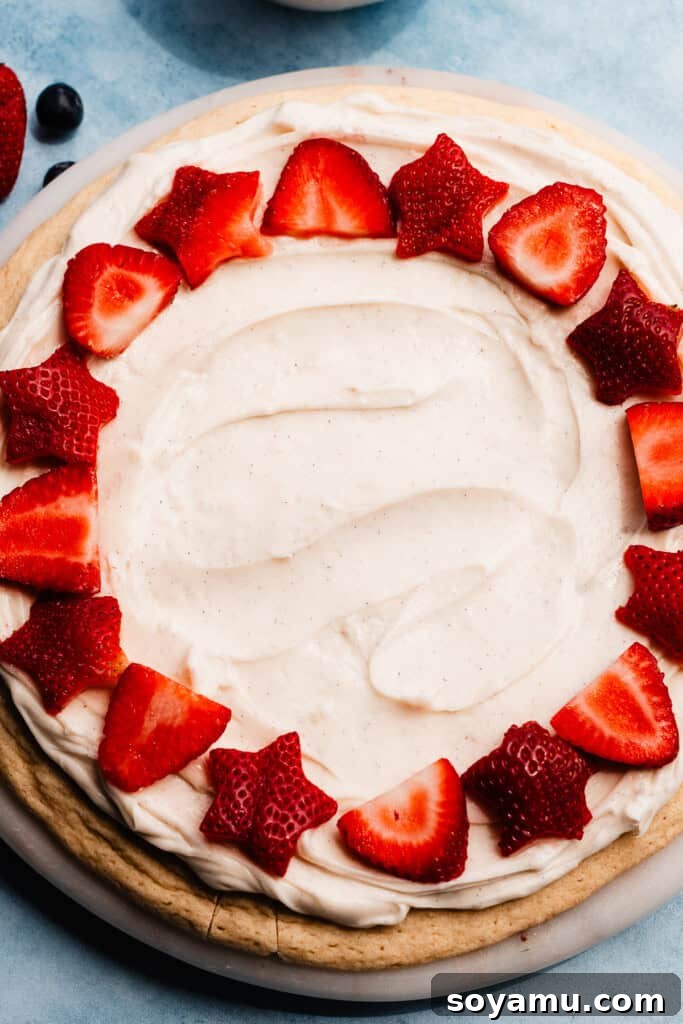 A close-up shot of a Fruit Pizza in the process of being decorated, with colorful fresh fruit being carefully arranged on top of the creamy white frosting and sugar cookie base.