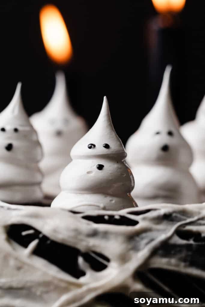 A close-up shot of two charming meringue ghosts, perfectly piped and decorated with expressive black gel frosting eyes and mouths, resting on top of the dark frosting of the Black Velvet Cake, ready for a Halloween celebration.