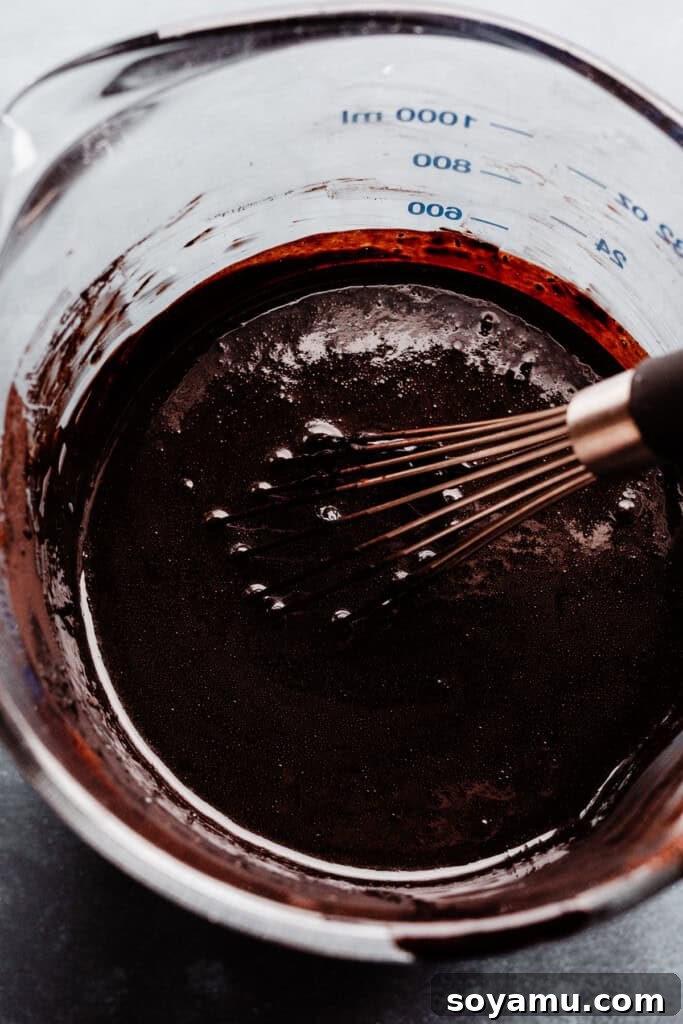 A close-up of bloomed black cocoa powder mixed with hot water in a glass measuring cup, creating an intensely dark liquid, ready to be incorporated into the cake batter for rich flavor and color.