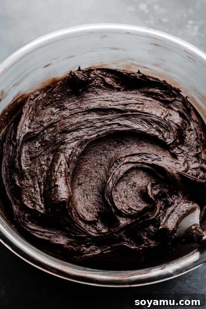 A generous bowl of rich, dark chocolate frosting, smooth and ready to be spread onto the cake layers. Its deep black color indicates the use of black cocoa powder.