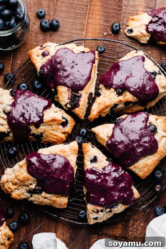 Double Blueberry Scones 11 Several golden-brown blueberry scones, freshly baked and glistening with a purple blueberry glaze, arranged on a rustic vintage wire cooling rack.