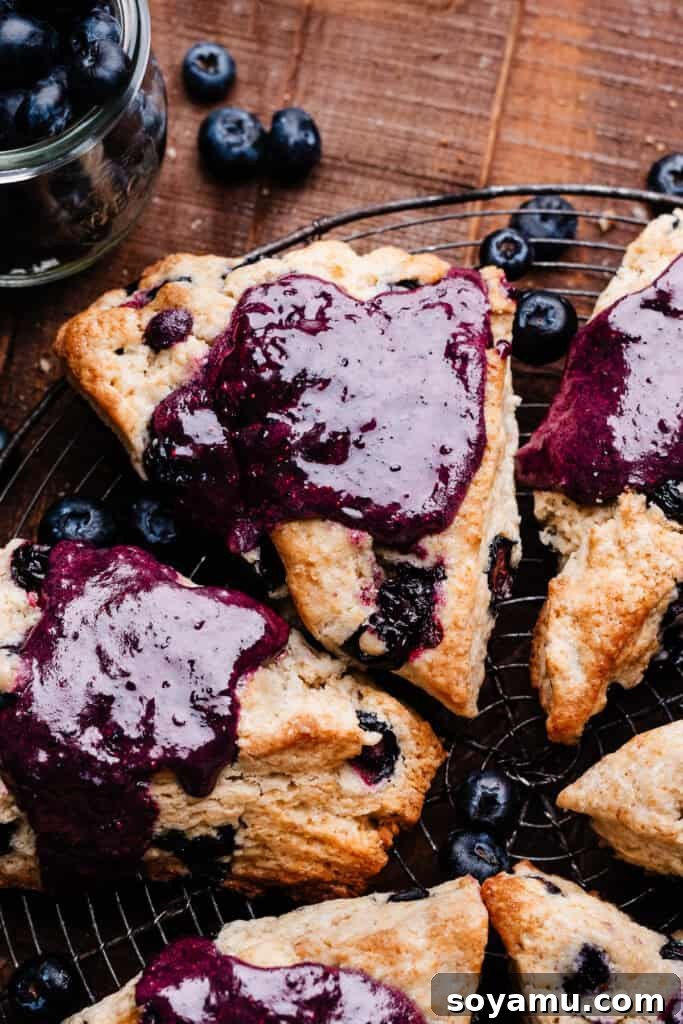 Double Blueberry Scones 3 A close-up shot of several freshly baked blueberry scones, beautifully glazed with a vibrant purple topping, resting on a rustic vintage wire cooling rack. The scones appear golden and inviting, ready to be enjoyed.