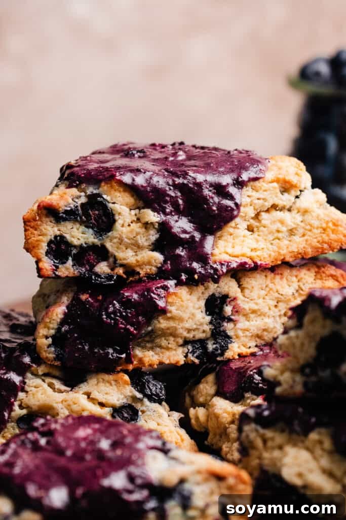 Double Blueberry Scones 10 A beautifully arranged stack of three golden-brown blueberry scones, each perfectly baked and generously drizzled with a rich purple glaze, showcasing their appealing texture and inviting presentation.