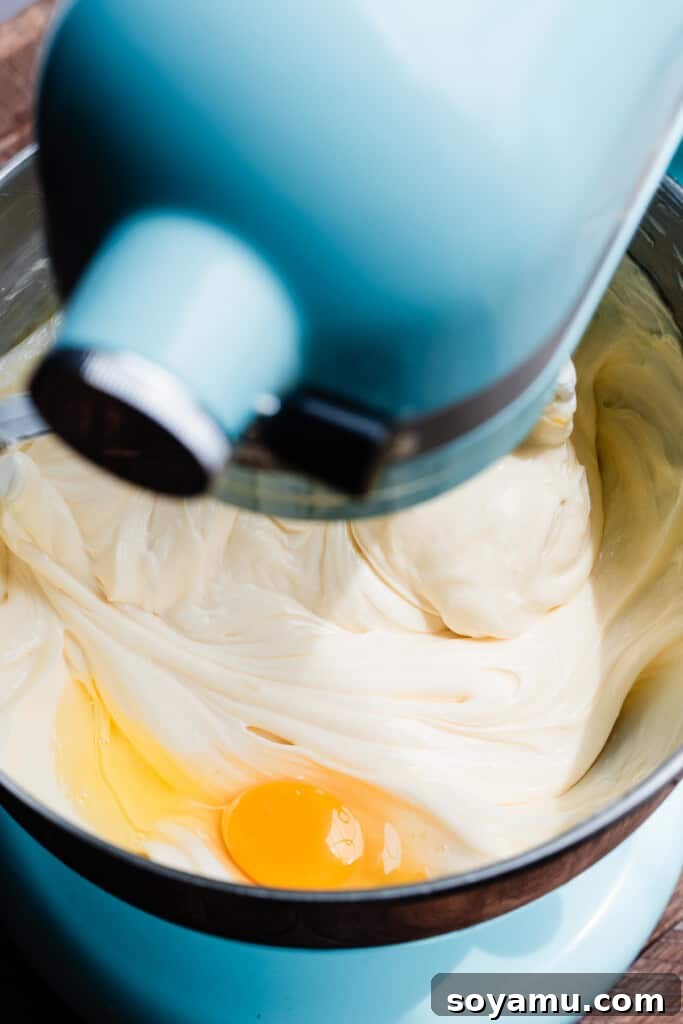 Velvety Rich New York Cheesecake Masterpiece 9 A fresh egg being cracked and added to the creamy cheesecake batter in a stand mixer, demonstrating the careful incorporation process.