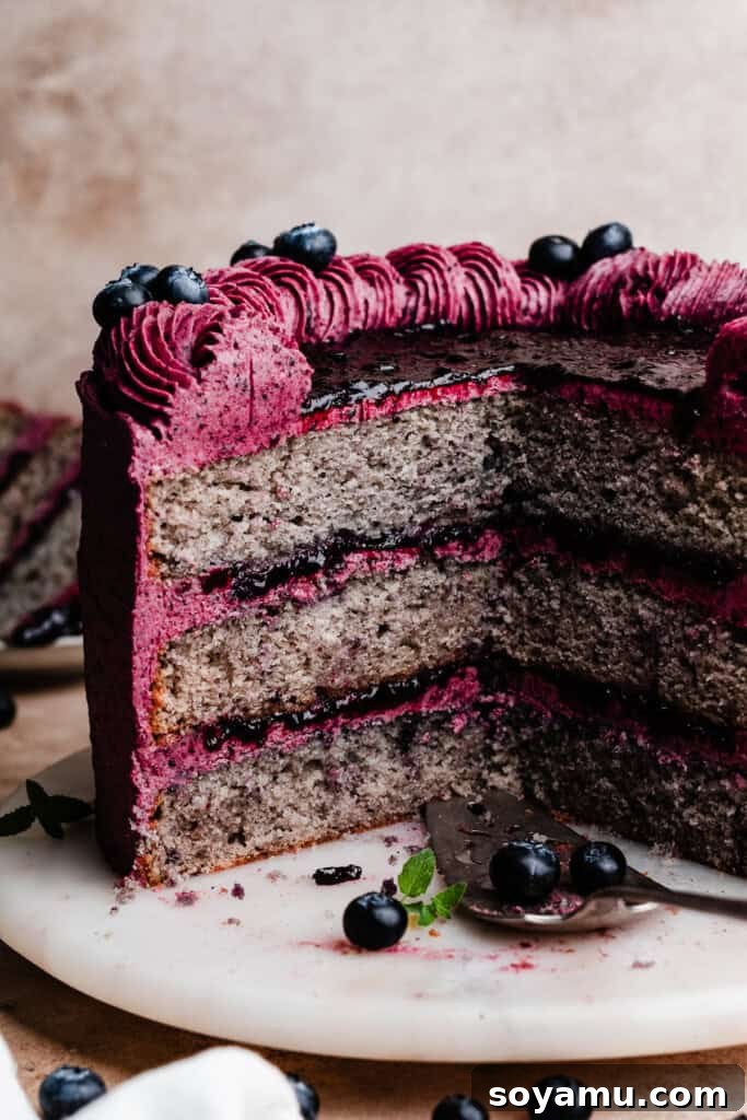 Blueberry Bliss Cake 2 A beautifully sliced blueberry cake on a white cake plate, showcasing its distinct layers of tender blueberry cake, vibrant blueberry compote filling, and smooth purple blueberry frosting.