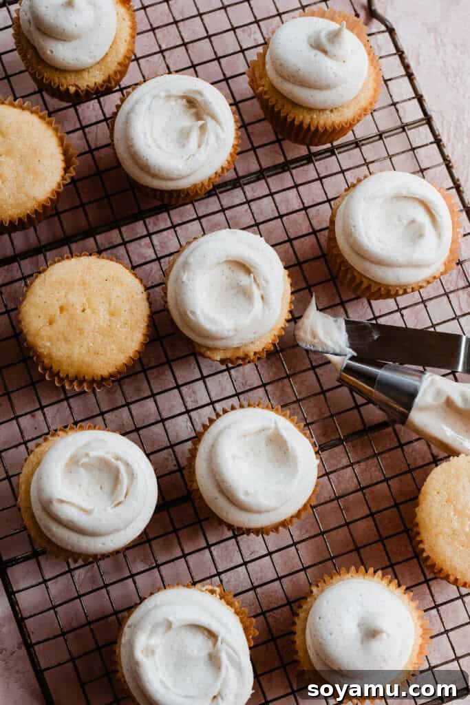 Several baked cupcakes resting on a wire rack, with one in the foreground being frosted with a generous swirl of vanilla buttercream.