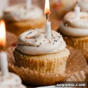 Vanilla cupcakes on a cake plate, topped with swirls of vanilla frosting, sprinkles, and lit birthday candles.