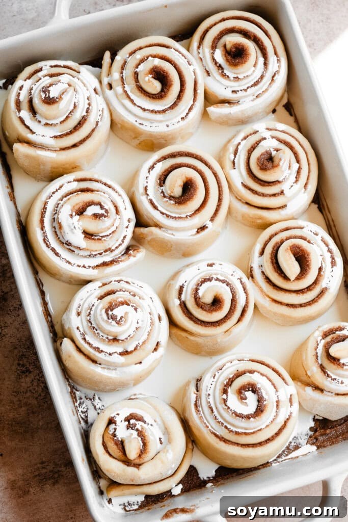 Heavy cream being poured generously over the risen cinnamon rolls in the baking pan, preparing them for baking.