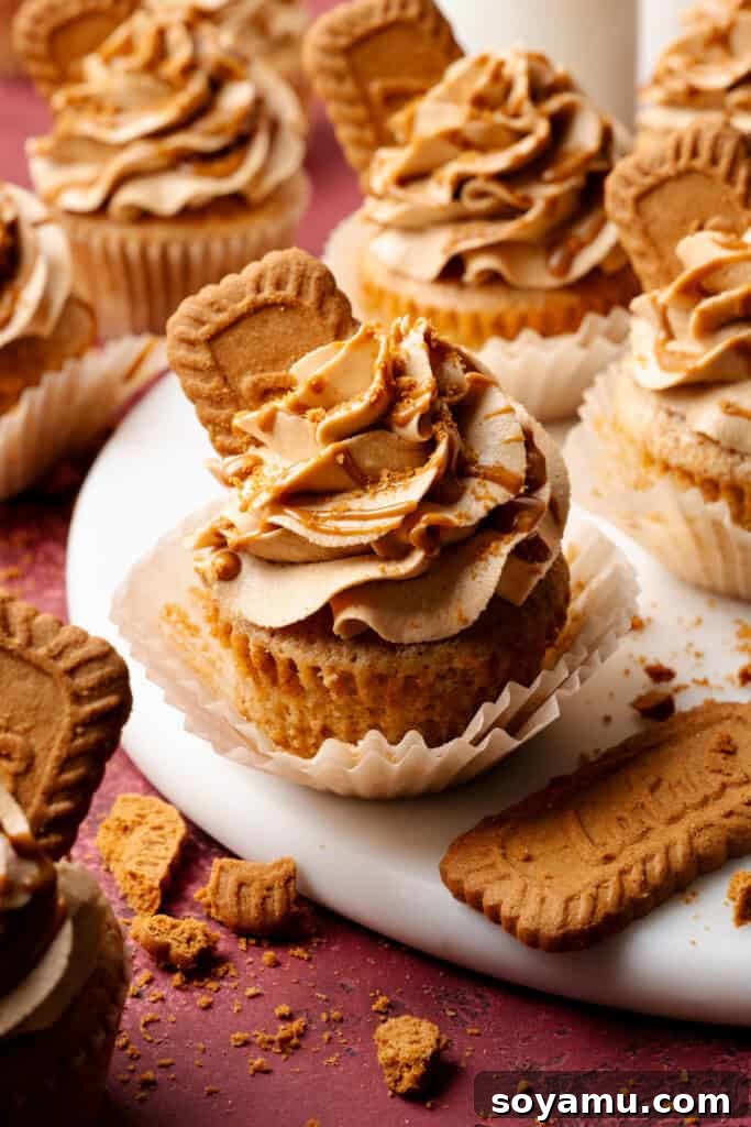 Biscoff Overload Cupcakes 2 A close-up of a biscoff cupcake on a marble cake plate. The cupcake is frosted with biscoff frosting, drizzled with cookie butter, and topped with half a biscoff cookie.