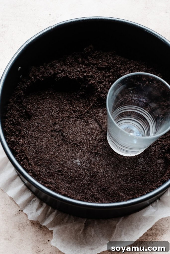 Simple Two Ingredient Oreo Dessert Crust 6 A hand using a juice glass to firmly press an Oreo crust mixture into a 9-inch springform pan, creating an even and compact base for a cheesecake.