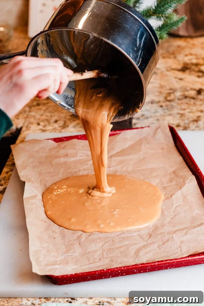 A stream of hot, golden toffee mixture being carefully poured from a saucepan onto a parchment-lined cookie sheet, ready for cooling and topping.