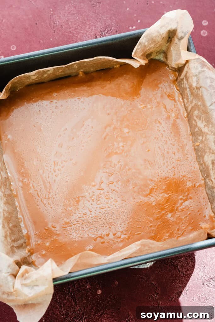 The molten toffee mixture evenly spread within a parchment-lined 9x9 inch square pan, showing the rich golden color before adding chocolate and nuts.