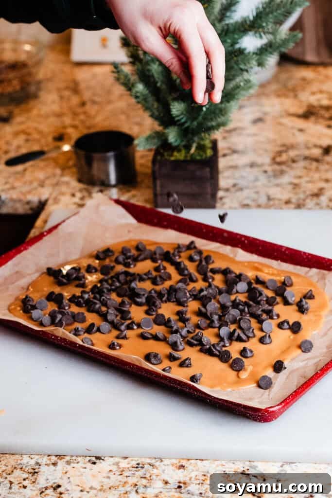 A hand sprinkling semi-sweet chocolate chips generously over the warm, golden toffee base on a parchment-lined cookie sheet, initiating the topping process.