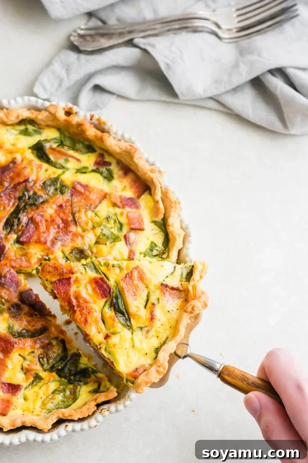 Perfect Breakfast Quiche - Our favorite Breakfast Quiche! The buttery flaky crust is filled with fluffy eggs, sharp cheddar cheese, spinach, and perfectly crispy bacon! #breakfastrecipes #easyrecipes #quiche #quicherecipes #baconrecipes #eggrecipes #mealideas | bluebowlrecipes.com A hand lifting a golden-brown slice of quiche, revealing its fluffy interior, out of the pie pan.