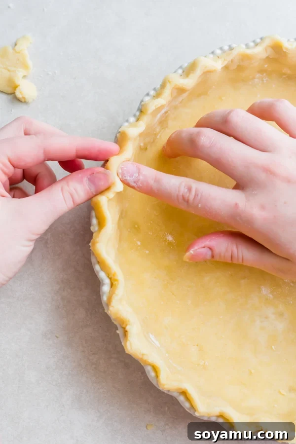 Perfect Breakfast Quiche - Our favorite Breakfast Quiche! The buttery flaky crust is filled with fluffy eggs, sharp cheddar cheese, spinach, and perfectly crispy bacon! #breakfastrecipes #easyrecipes #quiche #quicherecipes #baconrecipes #eggrecipes #mealideas | bluebowlrecipes.com Two hands meticulously crimping the edges of the pie dough to create a decorative, fluted crust.