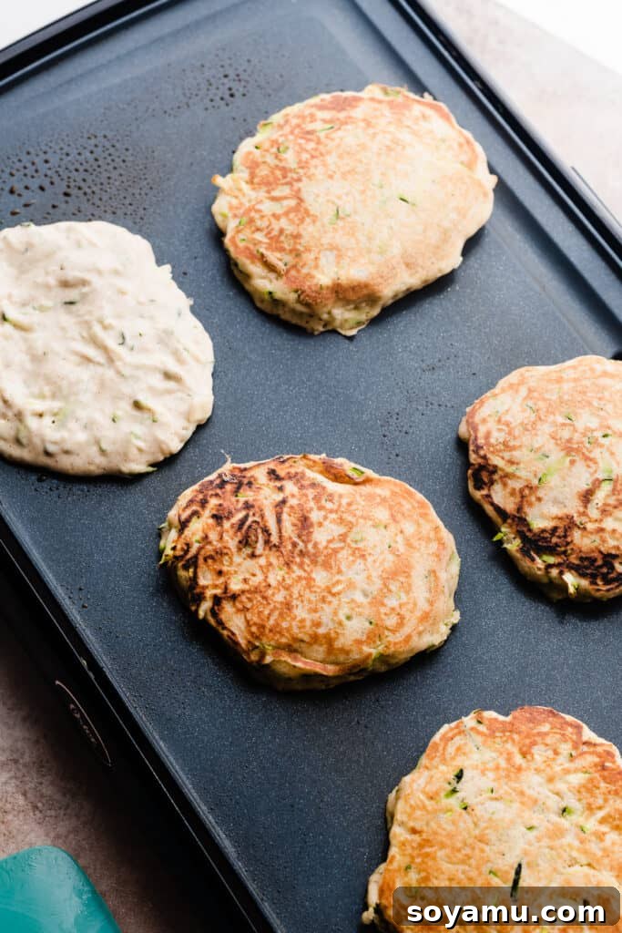Spiced Zucchini Pancakes 6 Zucchini pancakes cooking on a griddle, showing bubbles on the surface.