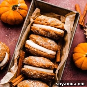 Frosted pumpkin whoopie pies in a stack on a burgundy backdrop, with mini pumpkins around, creating an inviting autumn scene.