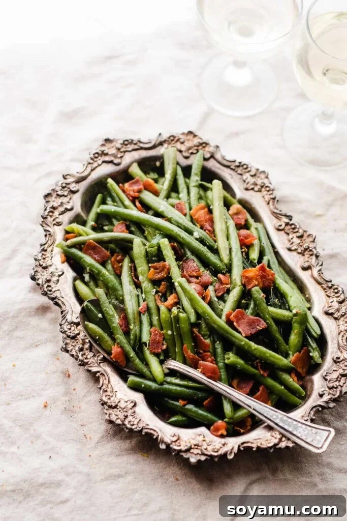 Effortless Gluten-Free Dairy-Free Bacon Green Beans 2 A metal dish of bacon green beans on a white linen surface, with a serving spoon.
