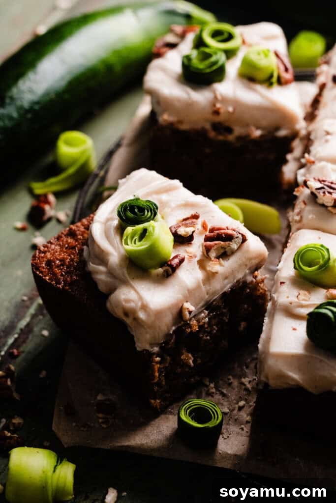 Irresistible Zucchini Snack Cake with Brown Butter Cream Cheese Frosting 9 A close-up of a delightful slice of zucchini cake on a white plate, adorned with frosting, chopped nuts, and delicate zucchini spirals, revealing its moist interior.