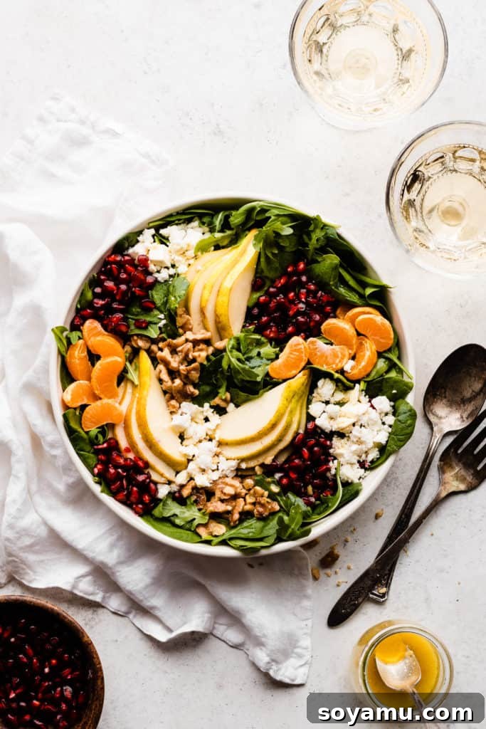 Jeweled Winter Pear Pomegranate Walnut Salad 6 A large salad bowl generously filled with a vibrant winter salad, showcasing a medley of fresh greens, artfully arranged sliced pears, clementine segments, glistening pomegranate arils, crumbled feta cheese, and whole walnuts.