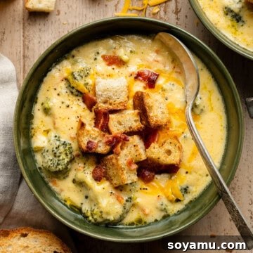 A close-up of a bowl of broccoli cheddar soup, topped with homemade croutons and bacon crumbles, looking incredibly appetizing.