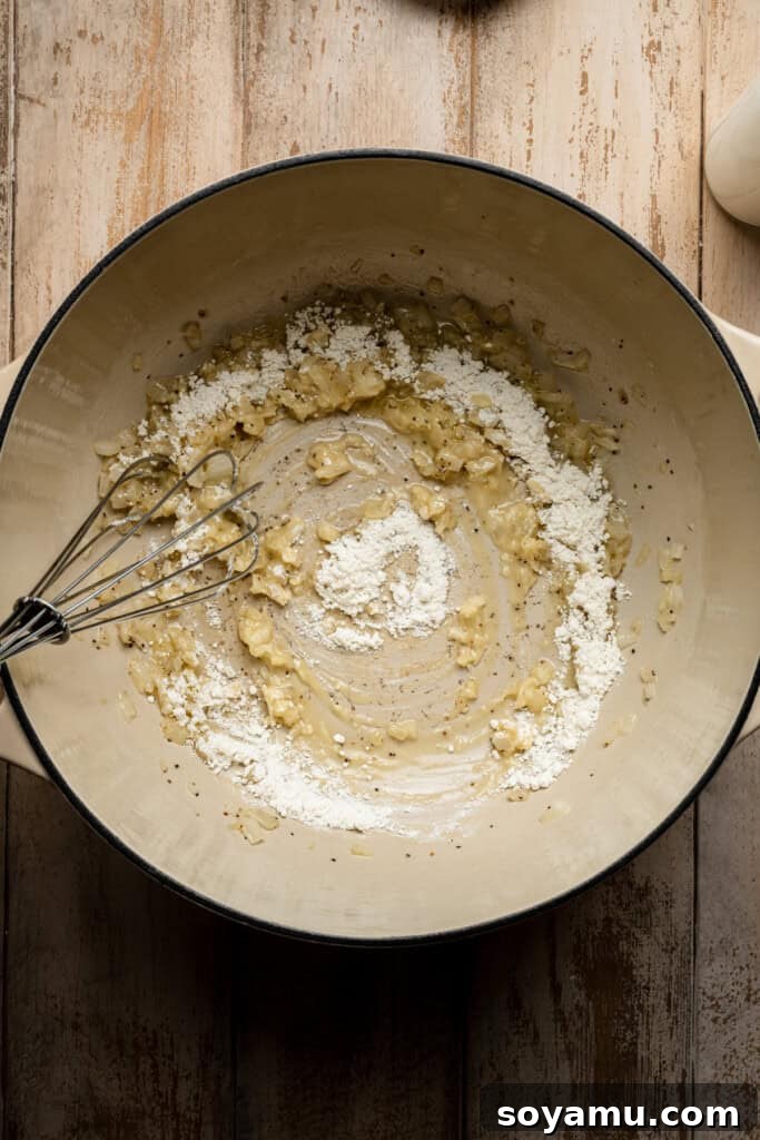A whisk vigorously mixing flour into the sautéed onions and garlic to form a roux, which will thicken the soup.